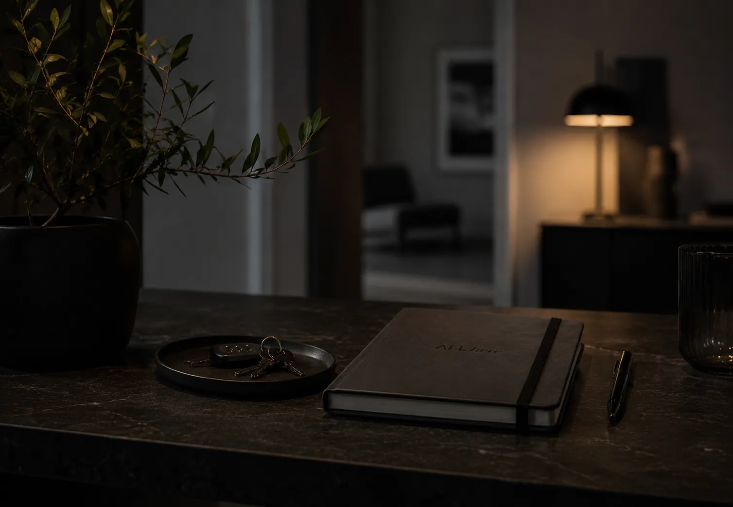 A debossed All Here notebook on a dark desk at dusk, with a small set of keys, an olive plant and a warm lamp in the background